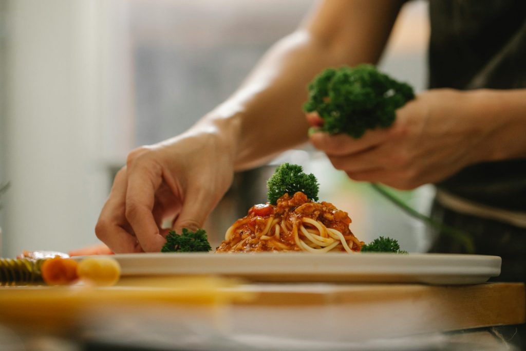 Crop anonymous housewife adding ripe parsley on delicious spaghetti with tomato sauce while cooking in kitchen